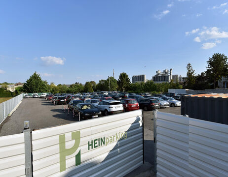 Parkplatz im Freien mit vielen geparkten Autos, einem Zaun im Vordergrund und Bäumen und Gebäuden im Hintergrund.