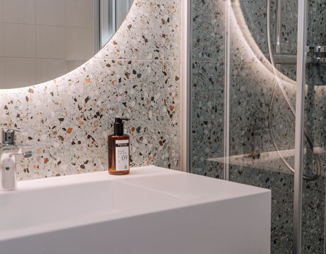 Modern bathroom with terrazzo wall, round, backlit mirror, white washbasin and brown soap dispenser.