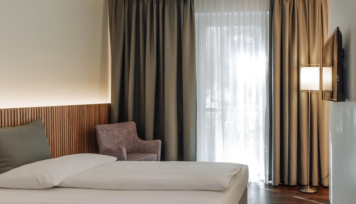 Minimalist hotel room with a bed, a gray chair, a floor lamp, a wall-mounted TV and a large window with curtains.