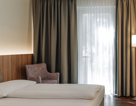 Minimalist hotel room with a bed, a gray chair, a floor lamp, a wall-mounted TV and a large window with curtains.