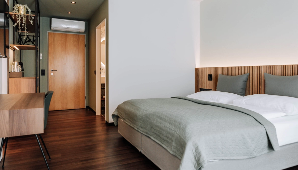 Modern hotel room with neatly made bed, wooden floor, desk and door to the bathroom.
