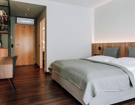 Modern hotel room with double bed, desk, wooden floor and a door to the bathroom.