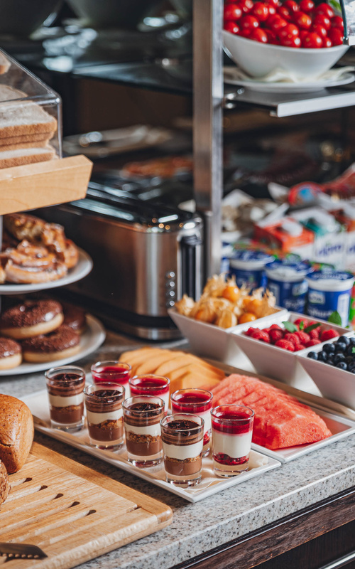 Frühstücksbuffet mit Gebäck, Obst, Miniparfaits, Joghurtbechern und einem Toaster auf einer Arbeitsplatte.