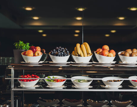 Weiße Schalen, gefüllt mit verschiedenen frischen Früchten und Gemüsesorten, die in Regalen auf einem Buffet präsentiert werden.