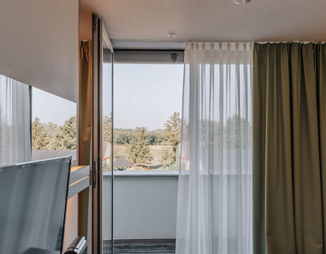 Open balcony door with sheer and green curtains, overlooking trees and rooftops under a clear sky.