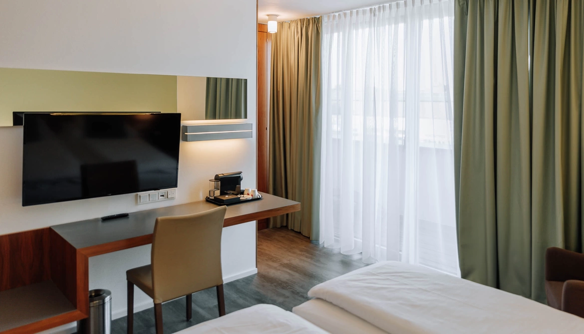 Modern hotel room with bed, desk, TV, chair, coffee machine and large window with green curtains.