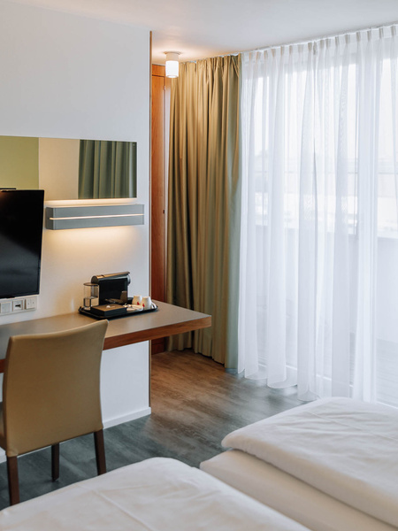 Modern hotel room with bed, desk, TV, chair, coffee machine and large window with green curtains.