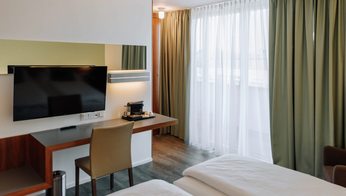 Modernes Hotelzimmer mit Bett, Schreibtisch, TV, Stuhl, Kaffeemaschine und großem Fenster mit grünen Vorhängen.