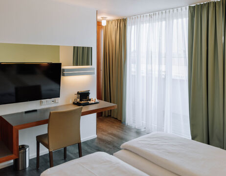 Modern hotel room with bed, desk, TV, chair, coffee machine and large window with green curtains.
