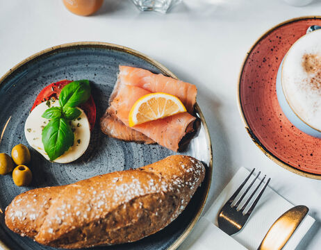 Teller mit geräuchertem Lachs, Zitrone, Mozzarella, Tomate, Oliven, Basilikum und Brot; Cappuccino als Beilage.