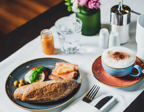 Frühstücksteller mit Brot, Mozzarella, Tomaten, Oliven, Schinken, Kaffee und Wasser auf einem gedeckten Tisch.