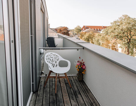 A small balcony with a modern white chair, wooden floor and a vase of colorful flowers on the railing.