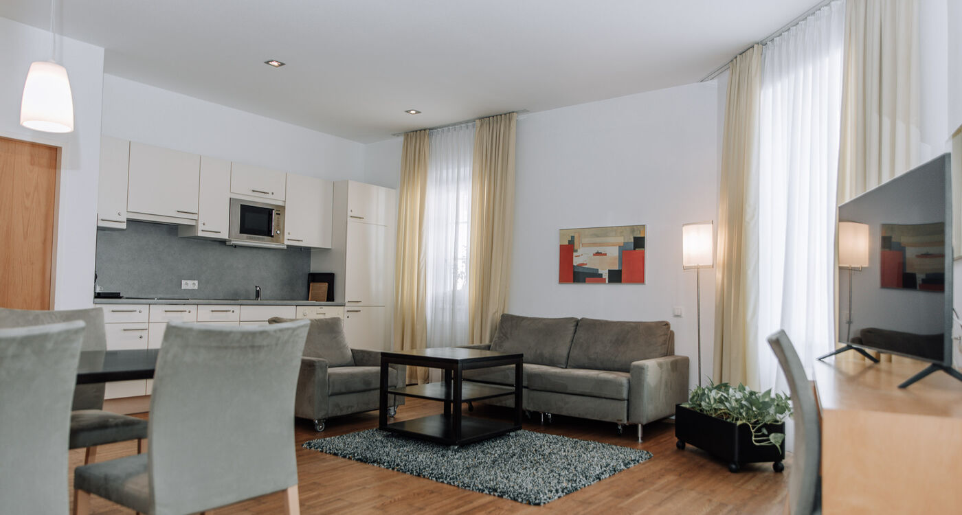 Modern living room with gray sofas, dining table, kitchenette, large windows and light curtains.