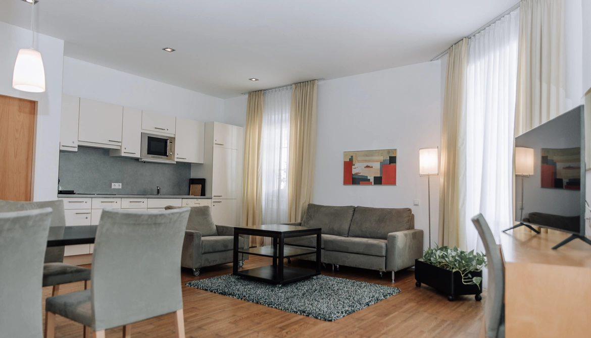 Modern living room with gray sofas, dining table, kitchenette, large windows and light curtains.