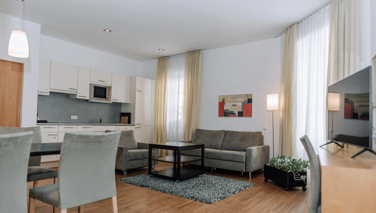 Modernes Wohnzimmer mit grauen Sofas, Esstisch, Küchenzeile, großen Fenstern und hellen Vorhängen.