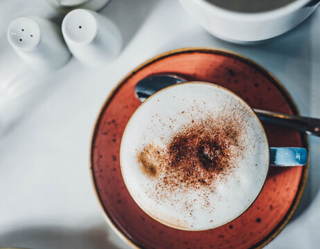 Tasse Cappuccino mit Zimt auf geschäumter Milch, auf einer roten Untertasse mit Löffel, von oben gesehen.