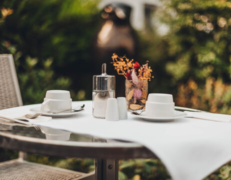 Tisch für zwei Personen im Freien mit weißen Tassen, Untertassen, Zuckerdose, Salz- und Pfefferstreuer und einer kleinen Blumenvase.