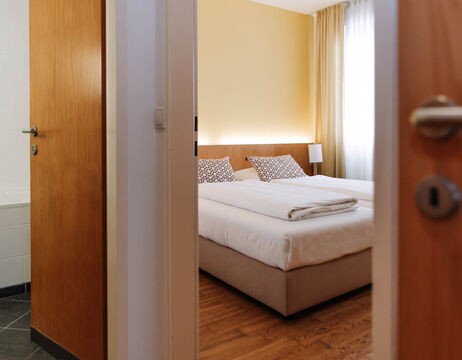 A neatly made bed with patterned cushions in a bright, modern bedroom, seen through an open door.