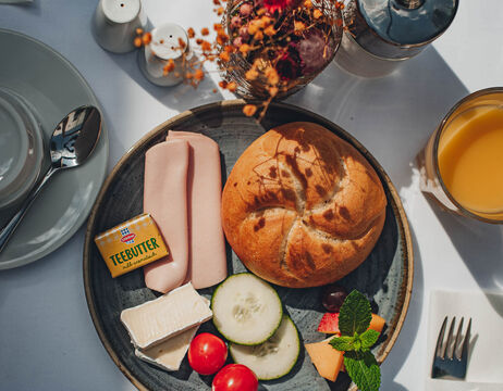 Ein Frühstücksteller mit Brot, Käse, Aufschnitt, Gurken, Tomaten, Butter und einem Glas Orangensaft.