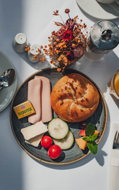 Place a plate with bread rolls, cheese, cold cuts, cucumber, tomato, butter and garnish on a laid table.