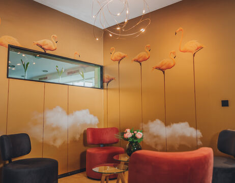 Lounge mit Flamingo- und Wolken-Wandkunst, roten und schwarzen Stühlen, Glastisch und Blumenvase.