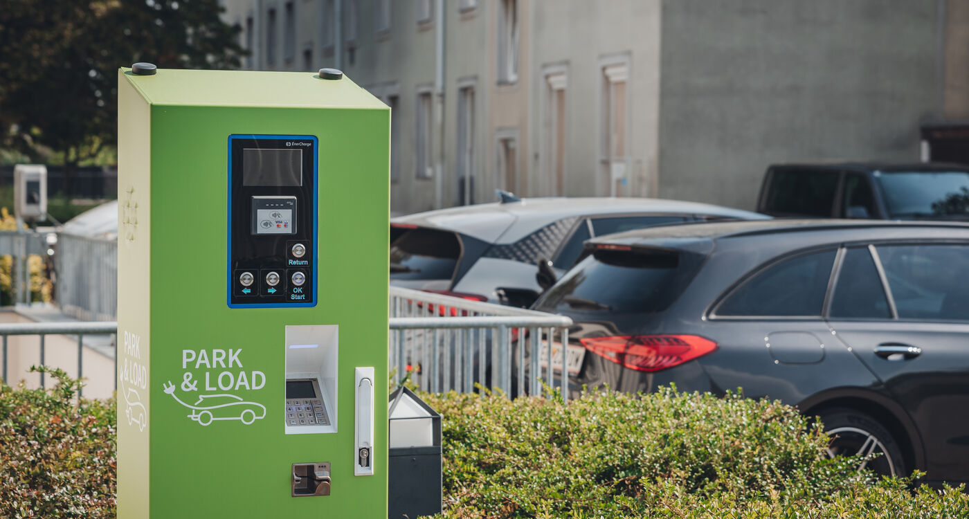 Grüne Ladestation für Elektrofahrzeuge in der Nähe geparkter Autos auf einem Parkplatz, mit Gebäuden im Hintergrund.