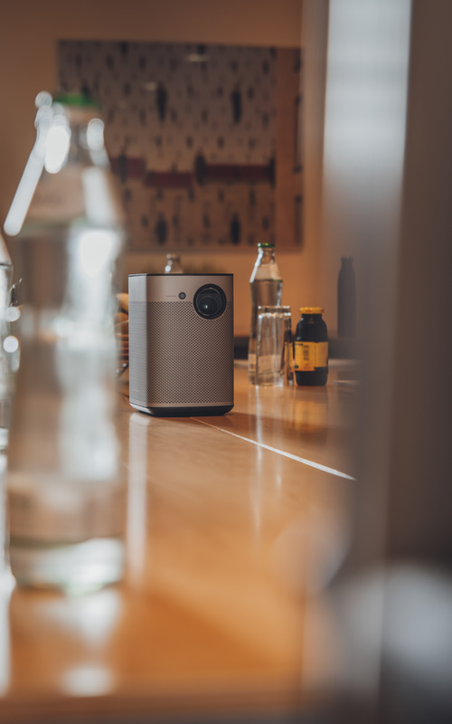 A portable projector stands on a wooden table surrounded by bottles in a dimly lit room.