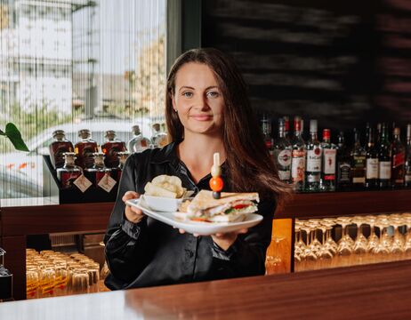 Eine Frau in einem schwarzen Hemd hält einen Teller mit einem Sandwich und Pommes an einer Theke.