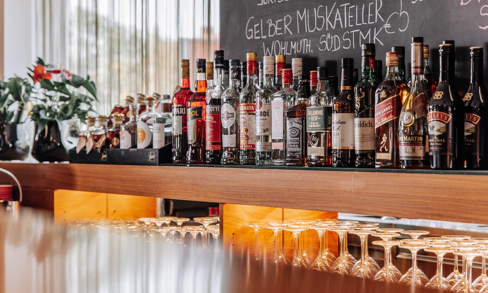 A row of different bottles of schnapps on a bar shelf with glasses underneath and a menu on a chalkboard above.