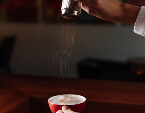 Eine Hand streut Zimt oder Kakaopulver auf eine Tasse Kaffee in einem roten Becher.