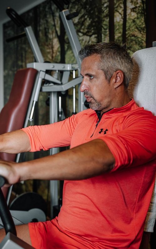 Mann in orangefarbener Sportkleidung bei der Benutzung von Fitnessgeräten in einem Fitnessstudio, mit einer Waldwand im Hintergrund.