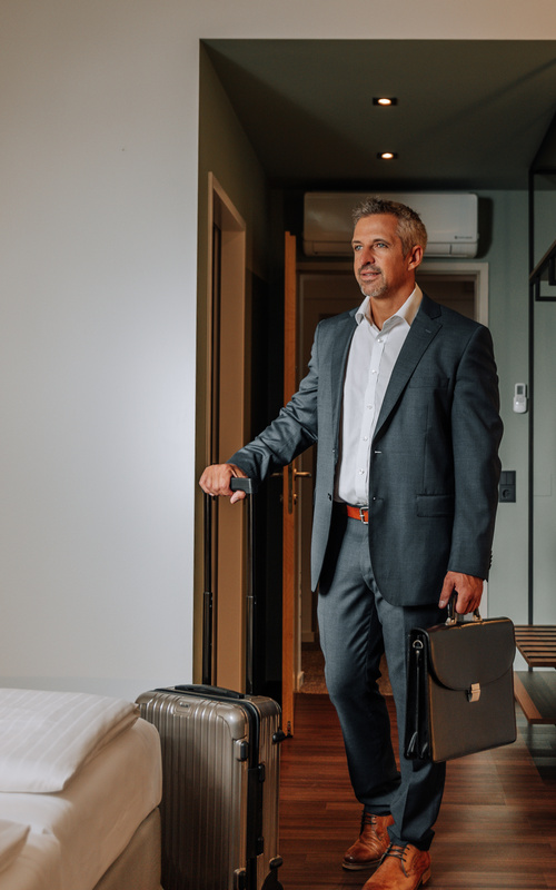 A businessman in a suit enters a hotel room, holding a suitcase and briefcase in his hand and looking ahead.