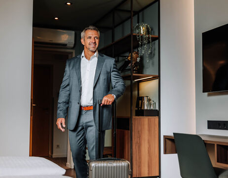 A smiling man in a suit enters a hotel room, dragging a suitcase behind him.