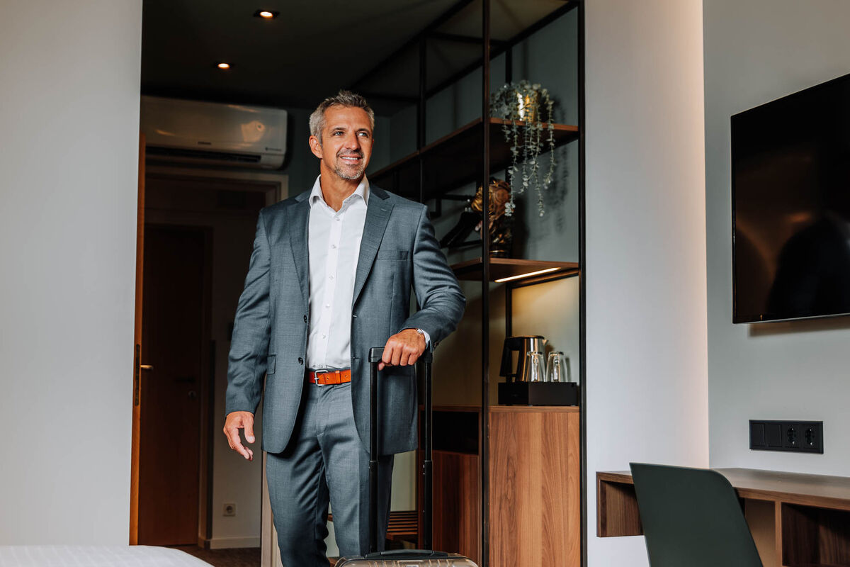 Ein lächelnder Mann im Anzug steht in einem modernen Hotelzimmer und hält den Griff eines Koffers.