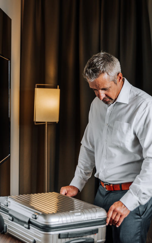 Man in a white shirt stands in a hotel room and packs a silver suitcase near a window with curtains.