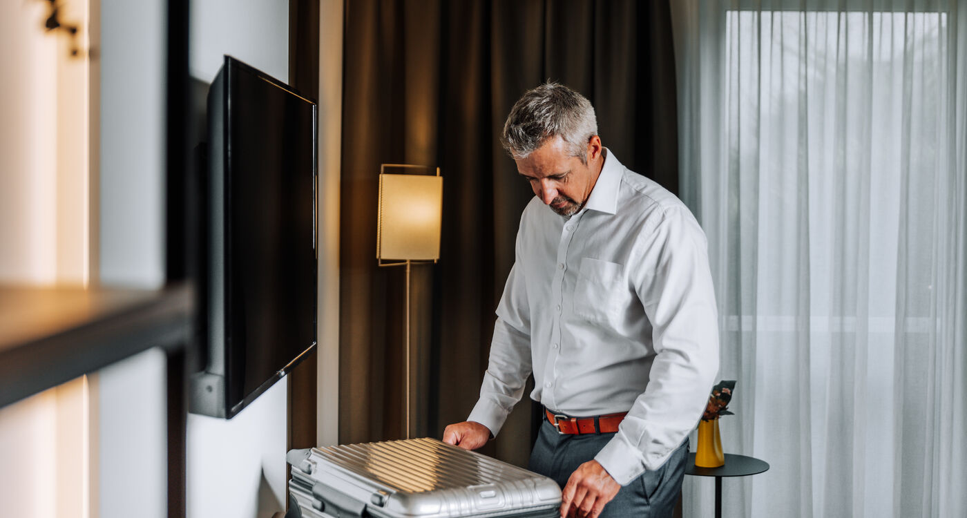 Man in a white shirt stands in a hotel room and packs a silver suitcase near a window with curtains.