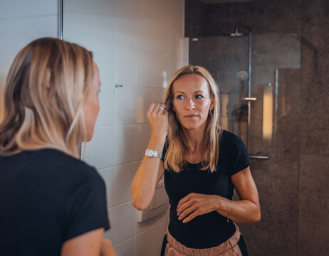 Frau, die sich vor einem Badezimmerspiegel schminkt, trägt ein schwarzes Hemd und eine Uhr.