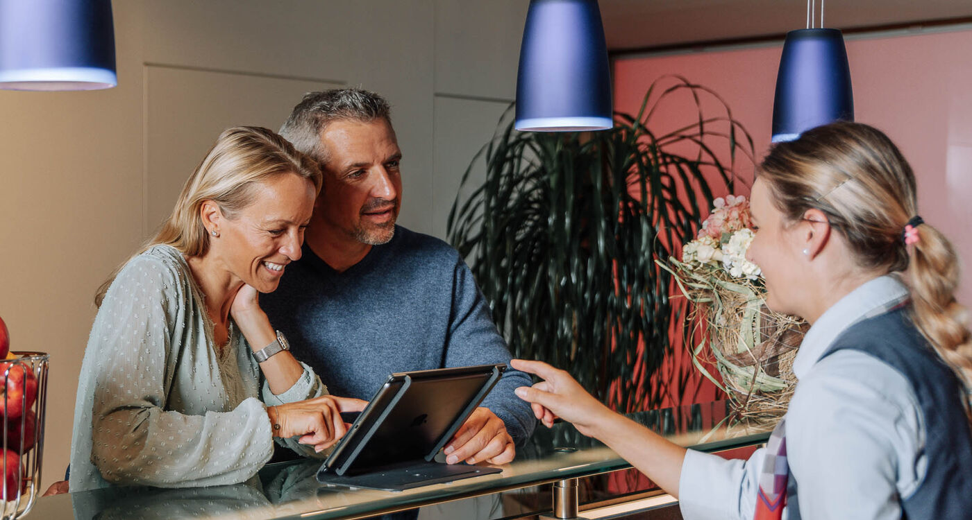 Ein lächelndes Paar checkt an der Rezeption ein, während die Rezeptionistin auf ein Tablet zeigt.