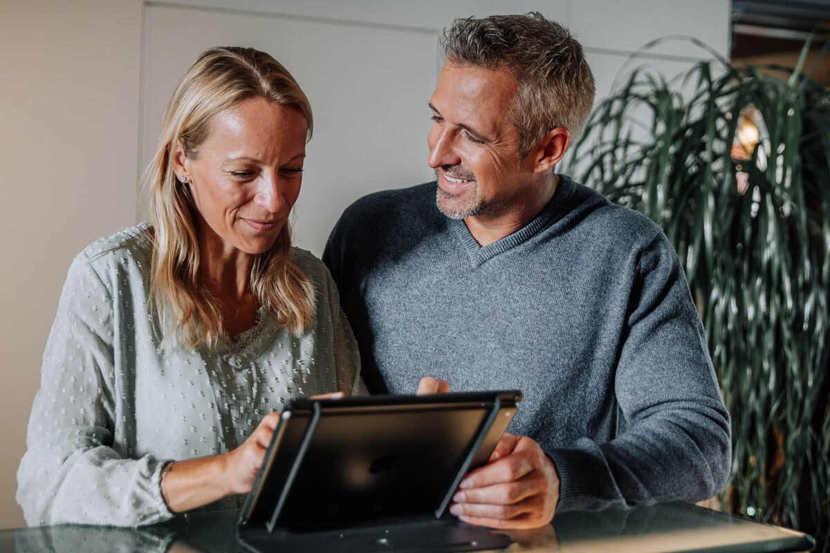 Ein lächelndes Paar sitzt zusammen und schaut auf ein Tablet, das auf einem Tisch in einem Haus steht.