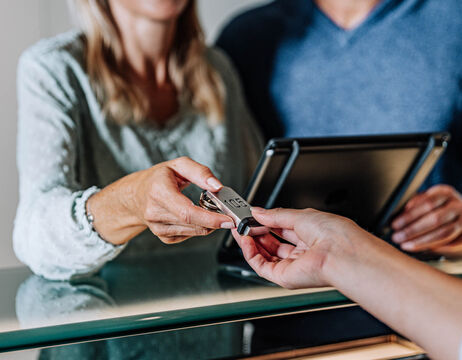 Eine Person übergibt einen USB-Stick an eine andere Person über einen Glastisch hinweg, mit einem Tablet im Hintergrund.