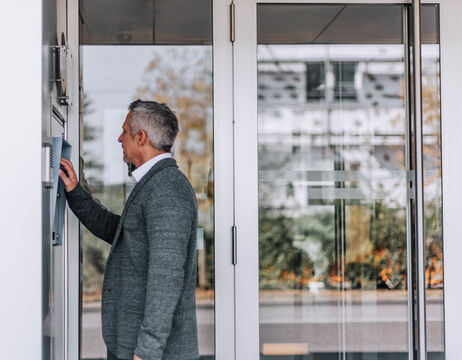 Ein Mann in einem grauen Pullover benutzt eine Gegensprechanlage vor einem Gebäude mit Glastür.