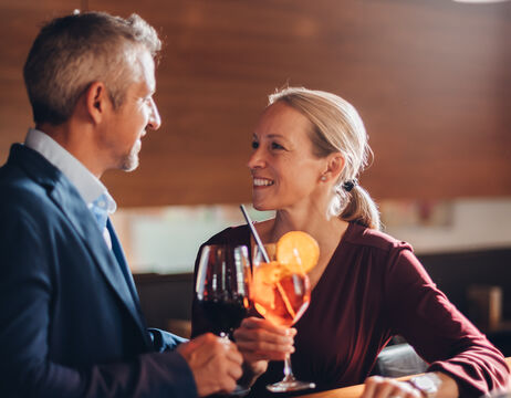 Ein lächelndes Paar sitzt an einer Bar, hält Getränke in der Hand und sieht sich liebevoll an.