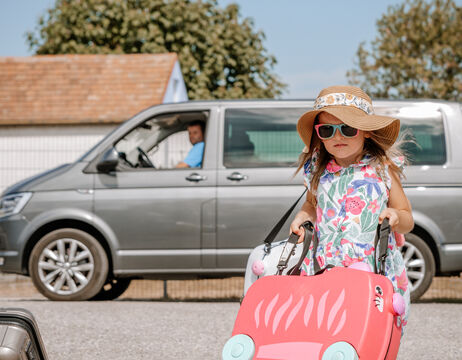 Ein junges Mädchen mit Sonnenbrille und Hut zieht einen rosafarbenen Koffer, während im Hintergrund ein grauer Lieferwagen mit Fahrer zu sehen ist.