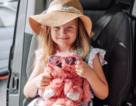 Junges Mädchen mit Sonnenhut, das lächelnd ein rosa Stofftier mit Sonnenbrille in einem Autositz hält.