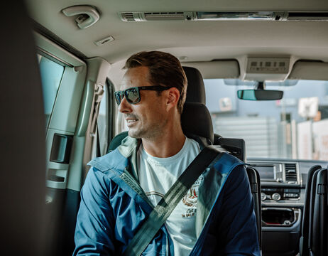 Ein Mann mit Sonnenbrille und Sicherheitsgurt sitzt auf dem Rücksitz eines Autos, schaut aus dem Fenster und lächelt.