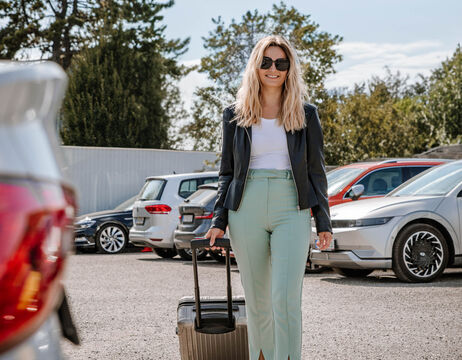 Frau mit blondem Haar und Sonnenbrille geht auf einem Parkplatz spazieren, zieht einen Koffer und lächelt.