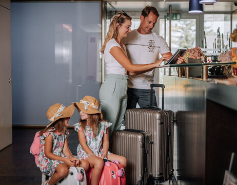 Eine Familie mit Gepäck checkt in einem Hotel ein; zwei junge Mädchen sitzen auf Koffern, die Eltern am Schalter.