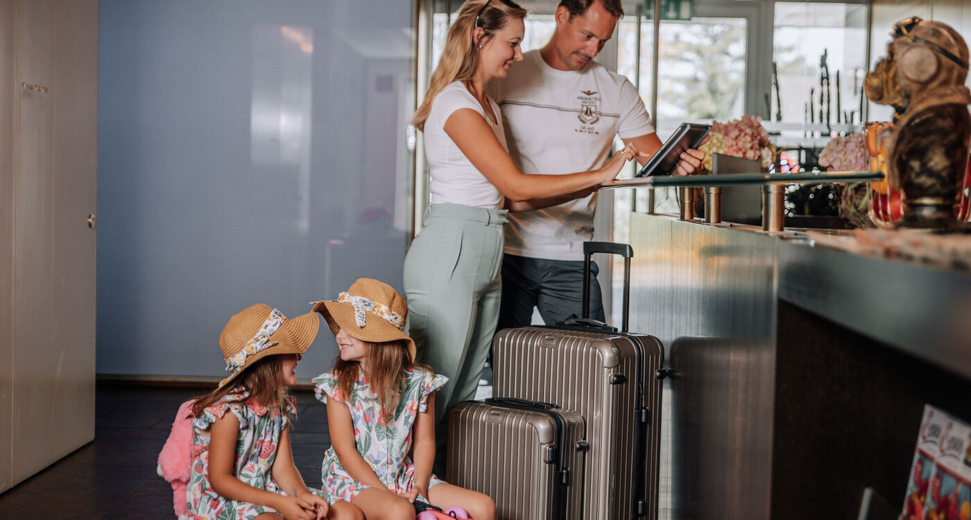Eine Familie checkt mit ihrem Gepäck an der Rezeption eines Hotels ein, während zwei junge Mädchen mit Hüten in der Nähe sitzen.