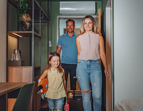 Eine lächelnde Familie mit Gepäck betritt ein Hotelzimmer, vor ihr eine Frau, ein Mann und ein kleines Mädchen.