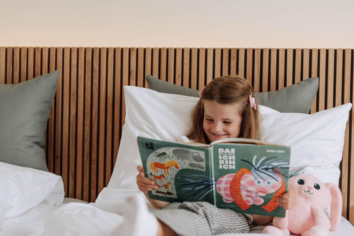 Ein lächelndes Mädchen liest im Bett ein Bilderbuch und hält ein rosa Stofftier neben sich.